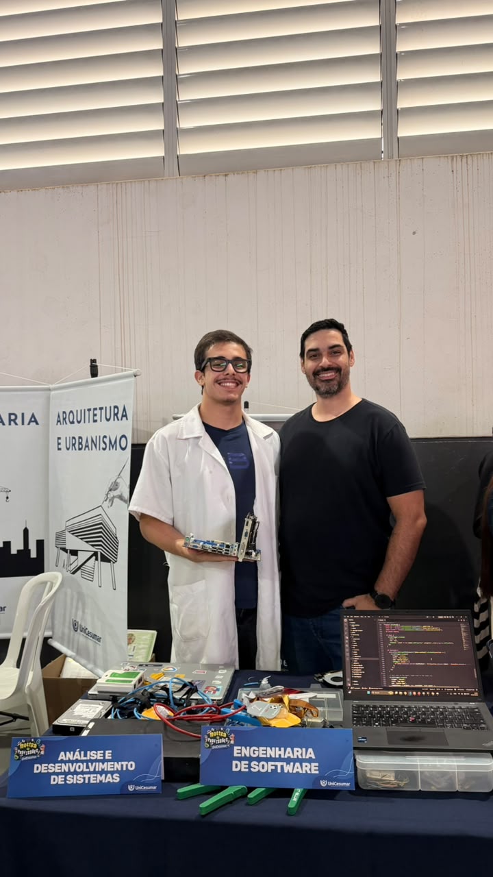 Dois integrantes da Axion atrás da mesa do estande, com as placas dos cursos de ADS e Engenharia de Software, componentes eletrônicos e um notebook exibindo código.
