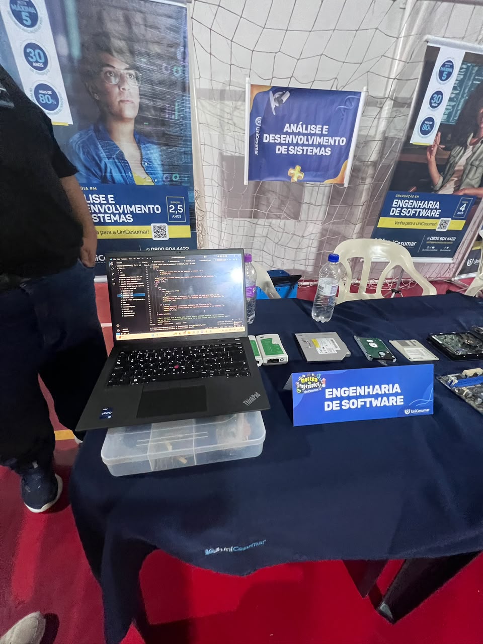 Estande da Axion em um ginásio, com notebook exibindo código, placa de Engenharia de Software e banners dos cursos de ADS e ESOFT ao fundo.
