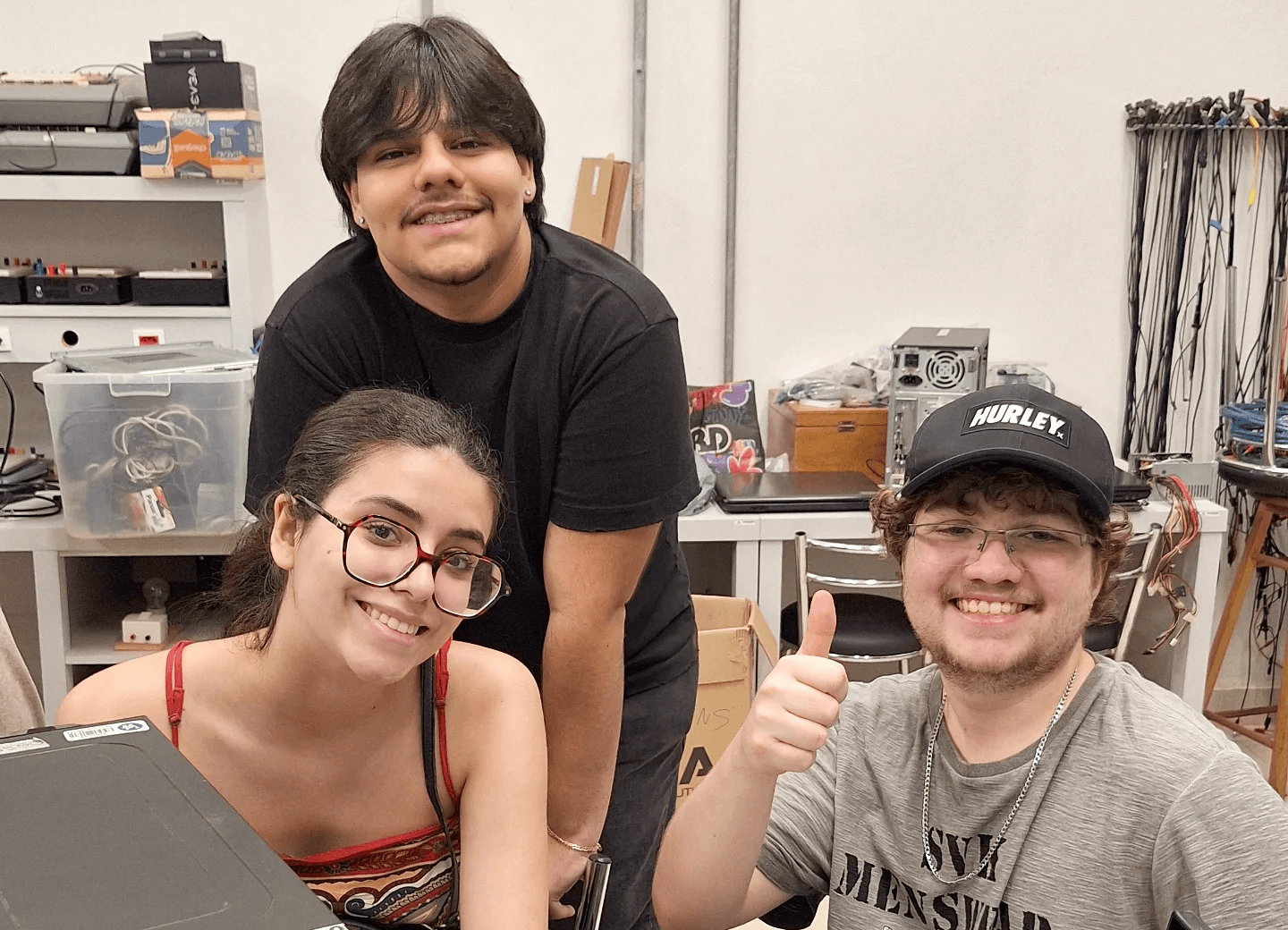 Três pessoas sorrindo em um laboratório de hardware, ao lado de um gabinete de computador.
