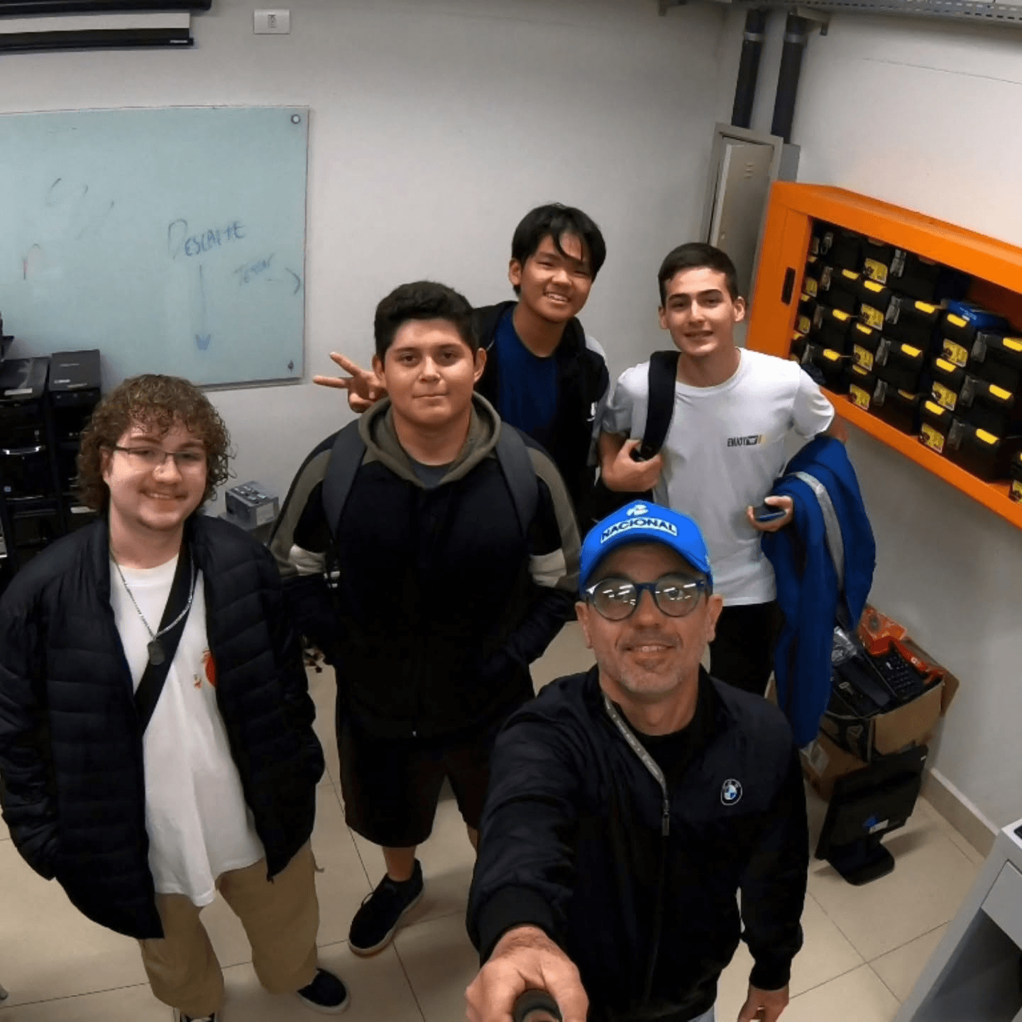 Grupo de pessoas em uma selfie no laboratório, com quadro branco e armário de ferramentas ao fundo.
