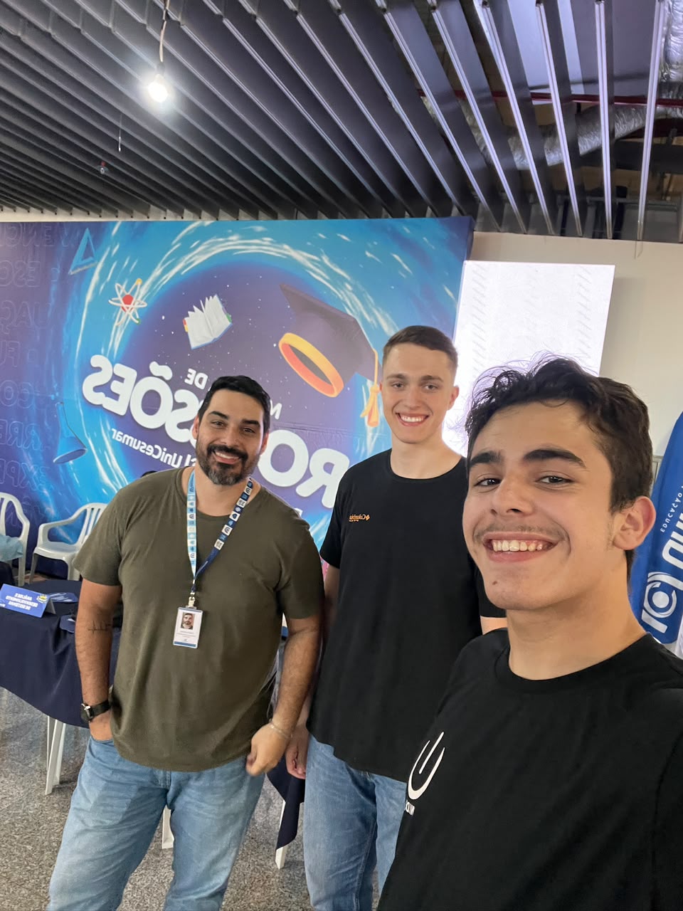 Três pessoas sorrindo em uma selfie diante do painel da Mostra de Profissões UniCesumar.